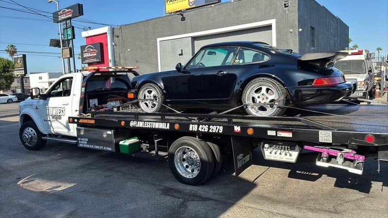 The Flawless Towing truck with a classic black Porsche 911 on the bed, company DOT and CA numbers visible